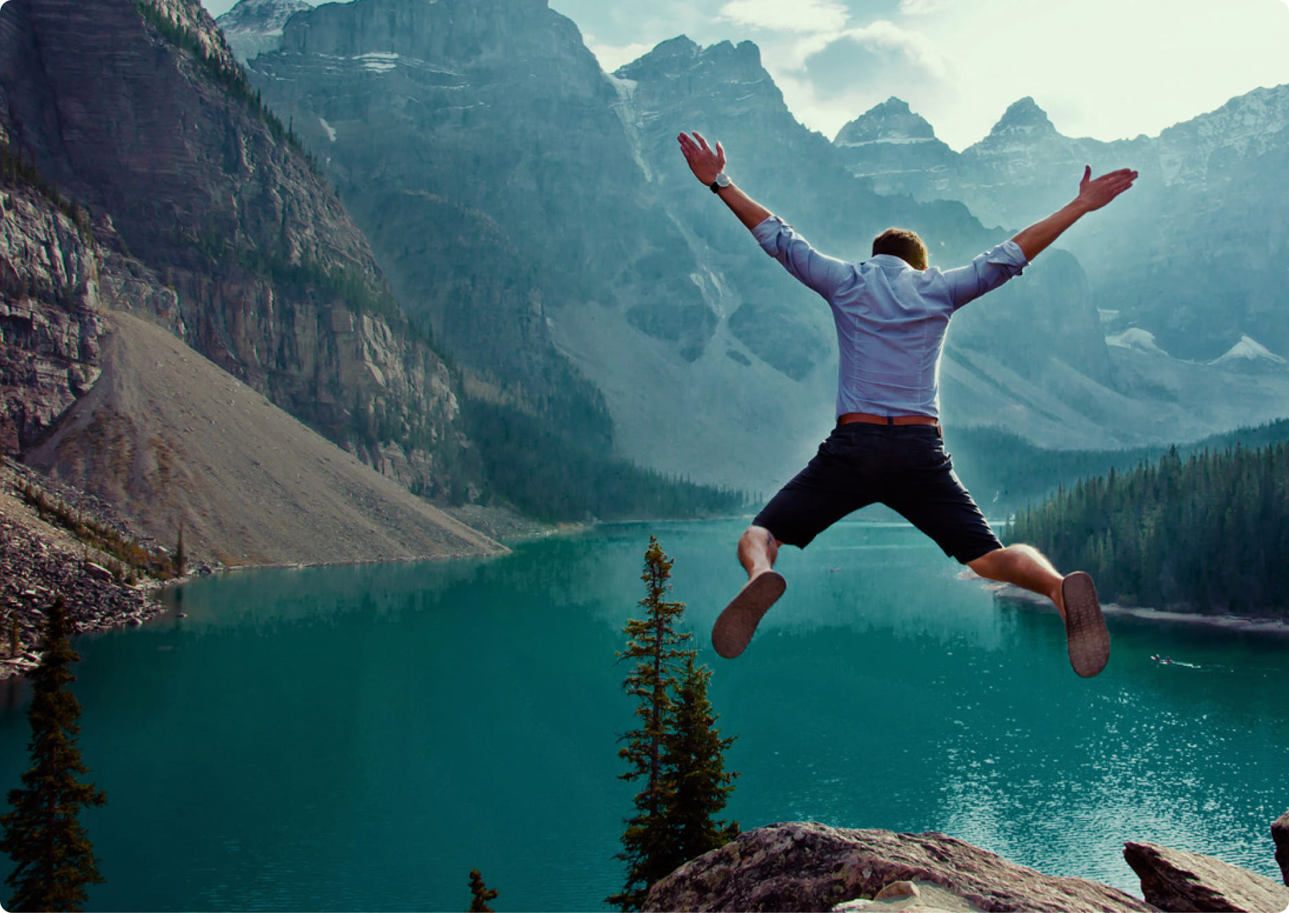 A person wearing a blue shirt and shorts jumps joyfully off a rock, overlooking a turquoise lake surrounded by pine trees and rugged mountain peaks under a cloudy sky.