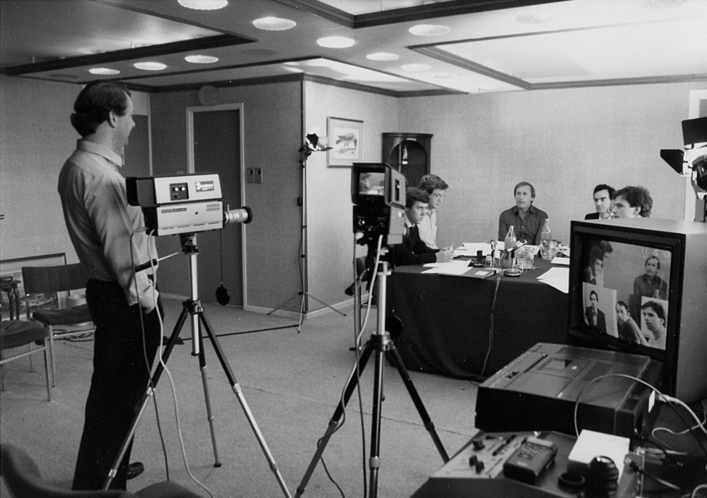 Black and white photo of a man operating a video camera pointed at a group of four people sitting at a table. Monitors and lights are set up around the room, showing a video feed of the group.