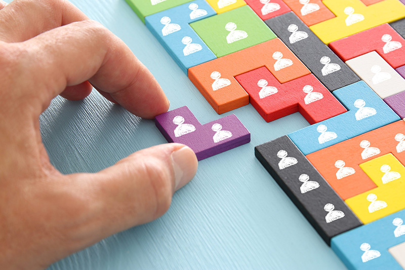 A hand places a purple puzzle piece among colorful, interlocking pieces, each with a white icon of a person, symbolizing teamwork or diversity in a team or organization.